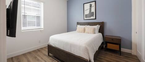 A bedroom with a bed, a nightstand, a wall-mounted TV, and a window with blinds.
