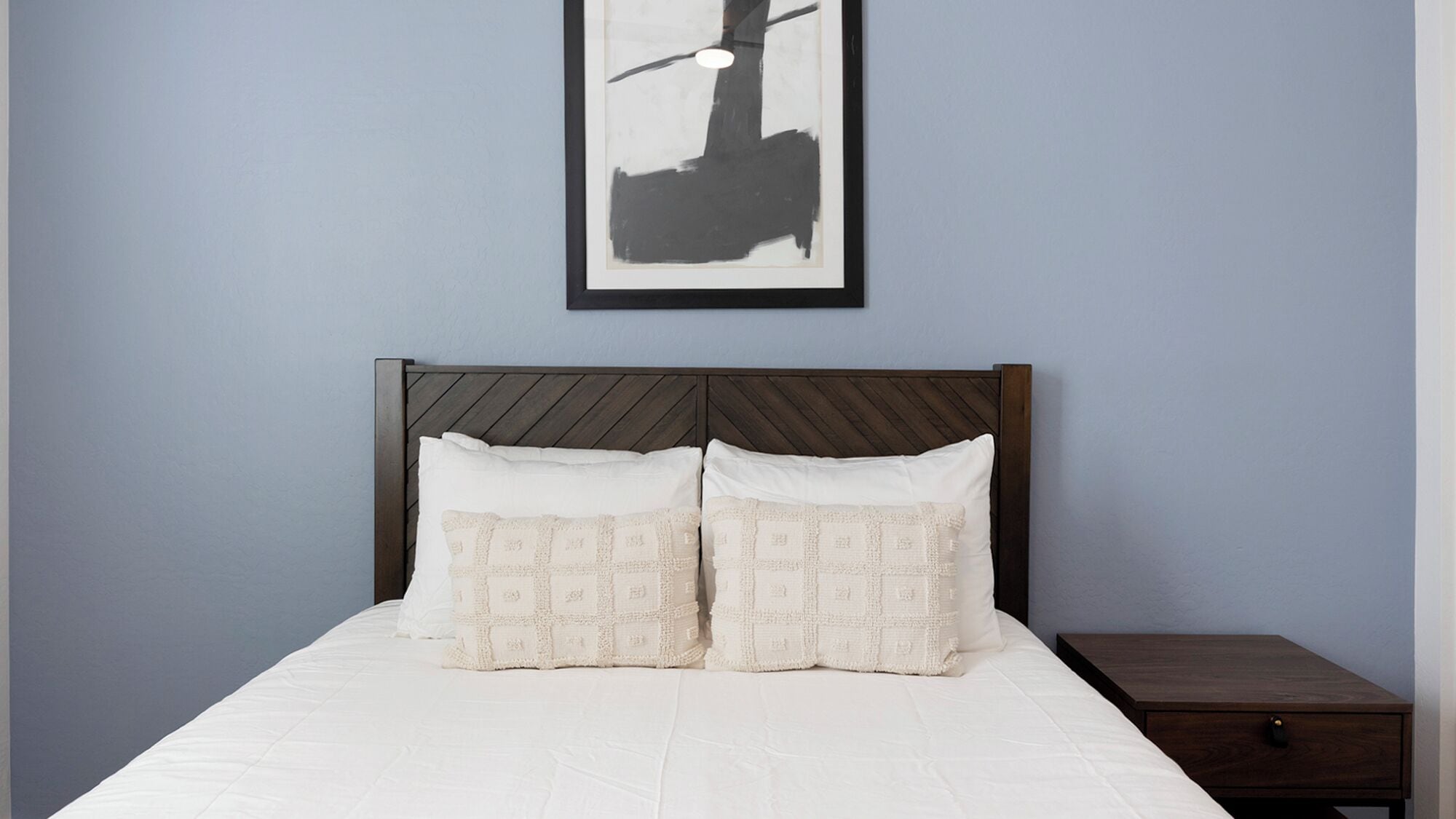A neatly made bed with two pillows, a wooden headboard, and a small wooden nightstand with a drawer.