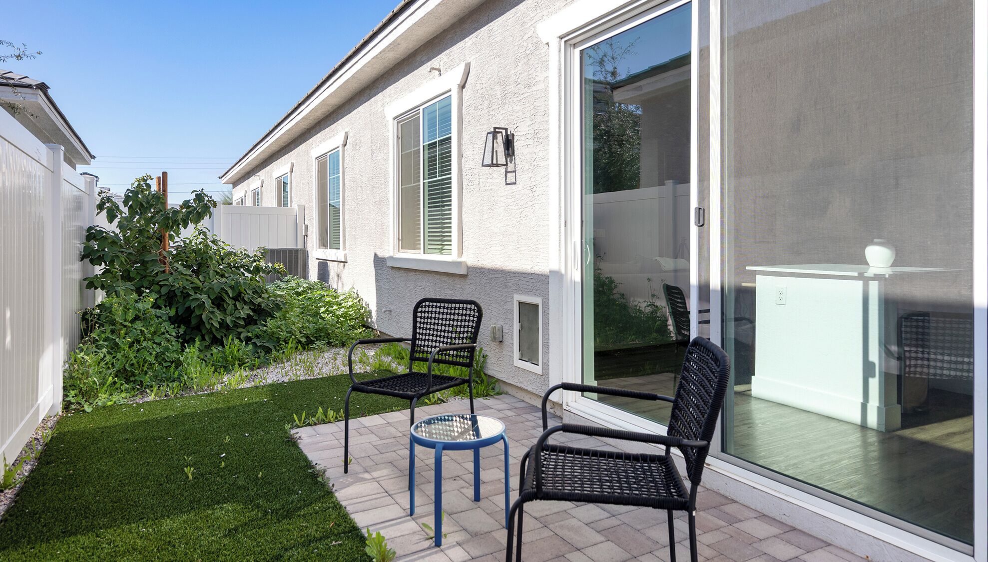 A patio area with two chairs and a small table, a white building with a sliding glass door, and a green lawn.