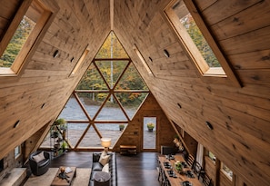 A wooden interior with a triangular skylight, a sofa, a dining table set, and a fireplace. - Timber & Glass: Elevated Luxe A-Frame • Hot-Tub • Fire-Pit • Nearby Creek Access (Mccaysville)