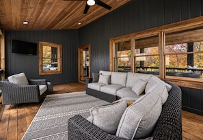 A room with a dark wooden ceiling, a large gray sectional sofa, a wicker chair, and a patterned rug. - Timber & Glass: Elevated Luxe A-Frame • Hot-Tub • Fire-Pit • Nearby Creek Access (Mccaysville)