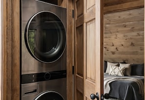 A laundry room with stacked washers and dryers, wooden paneling, and a view into a bedroom with a bed and pillows. - Timber & Glass: Elevated Luxe A-Frame • Hot-Tub • Fire-Pit • Nearby Creek Access (Mccaysville)
