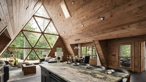 Ett rymligt rum i loftstil med ett stort träpanelat tak, en central köksö och stora fönster som erbjuder utsikt över den omgivande skogen.