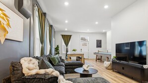 A modern living room with a sofa set, a coffee table, a TV, and a dining area with a table and chairs. - Luxury STL Oasis: Sauna, Fire pit, Hot Tub & Games (St. Louis)