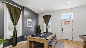 A modern room with a pool table, dark grey walls, and a door with a window view. - Luxury STL Oasis: Sauna, Fire pit, Hot Tub & Games (St. Louis)