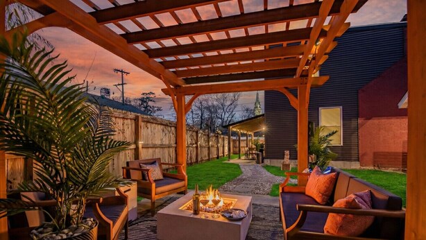 A covered outdoor seating area with a fire pit, wooden furniture, and a view of a house in the background. - Luxury STL Oasis: Sauna, Fire pit, Hot Tub & Games (St. Louis)