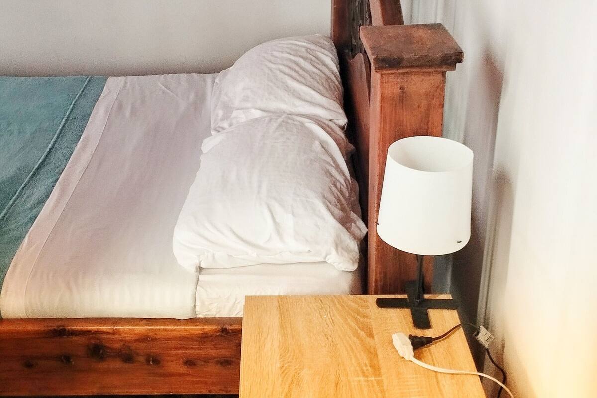 A small, single-bed room with a wooden headboard, a bedside table with a lamp, and a wooden floor.
