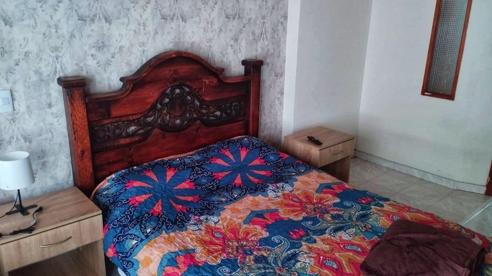Ein Schlafzimmer mit einem hölzernen Kopfteil, einer blumigen Tagesdecke, einem Nachttisch mit Lampe und einem kleinen Beistelltisch.