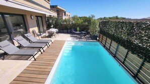 Una piscina en la azotea con sillones de descanso y una mesa, rodeada de follaje y un edificio al fondo.