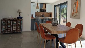Una cocina moderna con encimera de madera y luces colgantes, un comedor con mesa de madera y sillas naranjas, y un amplio ventanal con vistas al exterior.