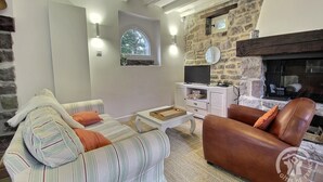 Ein gemütliches Wohnzimmer mit Kamin aus Stein, gestreifter Couch, Ledersessel und Fernseher auf einem weißen Schrank.