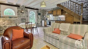 Ein gemütliches Wohnzimmer mit einem Ledersessel, einem gestreiften Sofa und einem Esstisch mit Stühlen. Im Hintergrund ist eine Küche mit Holzschränken und einer Steinwand zu sehen.