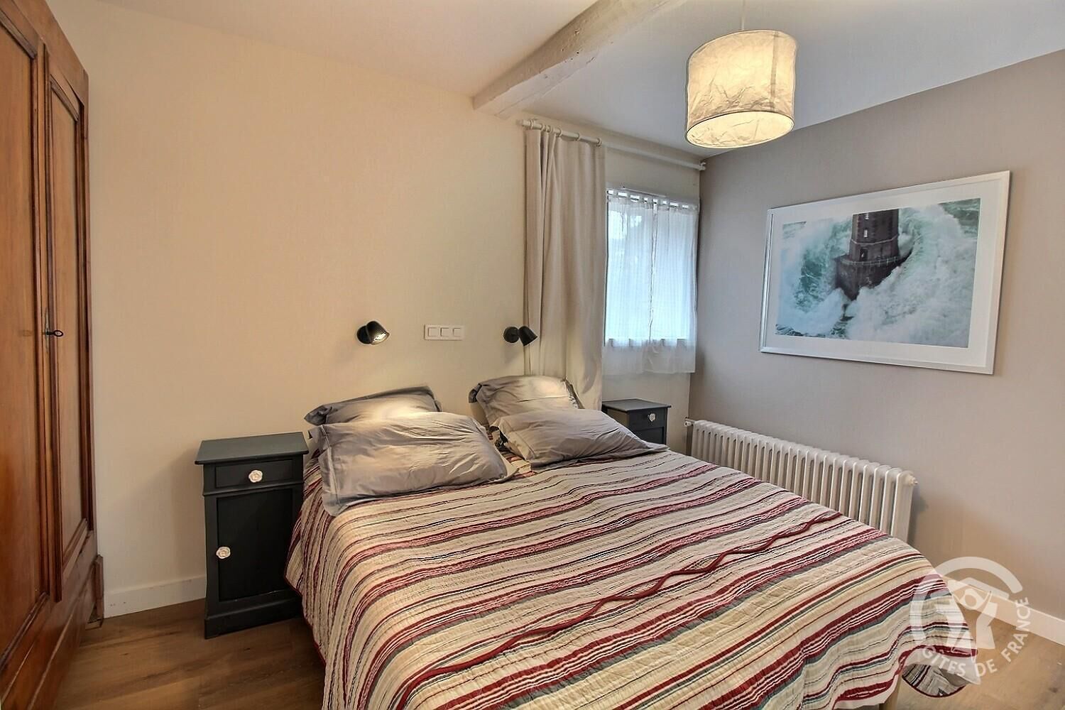 Une chambre à coucher avec un lit, des tables de chevet, un radiateur, une fenêtre avec des rideaux et un tableau au mur.