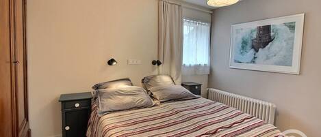 Une chambre à coucher avec un lit, des tables de chevet, un radiateur, une fenêtre avec des rideaux et un tableau au mur.