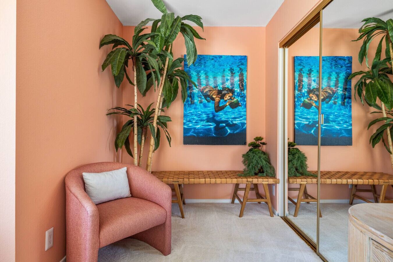 Una habitación con una silla rosa, dos plantas de bambú, una mesita y dos cuadros grandes de una persona nadando.