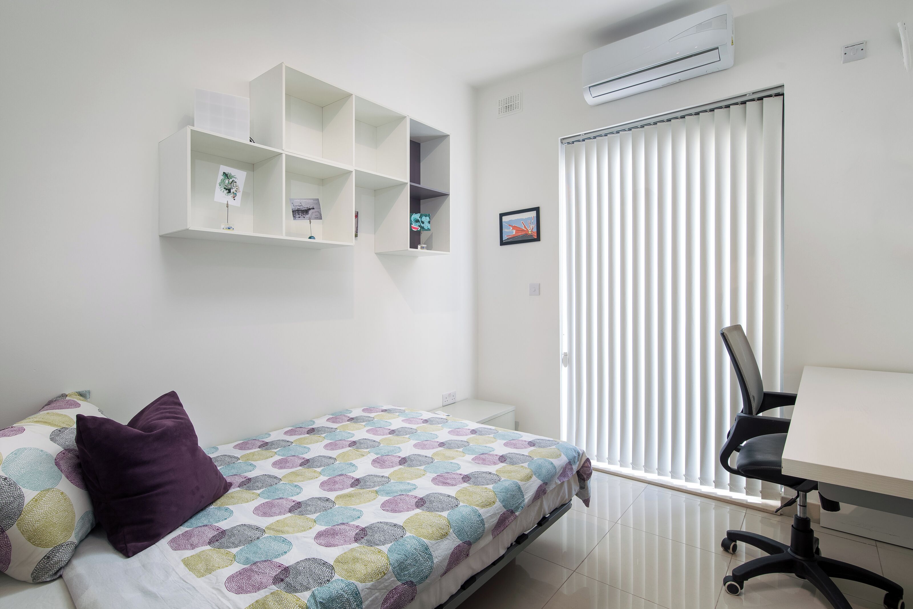 Una camera da letto con un letto, una scrivania e una sedia. Si vede una finestra con persiane, un condizionatore d'aria e uno scaffale con oggetti appoggiati al muro.