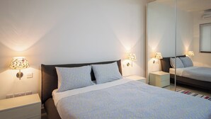 Ein ordentlich bezogenes Bett mit einem gemusterten Überwurf, zwei Nachttischlampen, einem Nachttisch und einem großen Spiegel an der Wand.