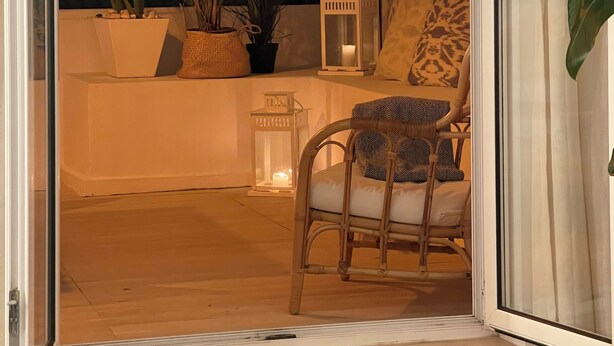 Eine geöffnete Schiebeglastür führt zu einem Balkon mit einem Korbstuhl, einem kleinen Tisch mit Kerze und Topfpflanzen.