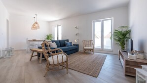 Ein geräumiges Wohnzimmer mit einem blauen Sofa, Korbstühlen, einem Esstisch und großen Fenstern.