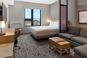 Une chambre d’hôtel comprenant un lit, un bureau, une chaise, un canapé, une petite table en bois et une grande fenêtre donnant sur la ville.