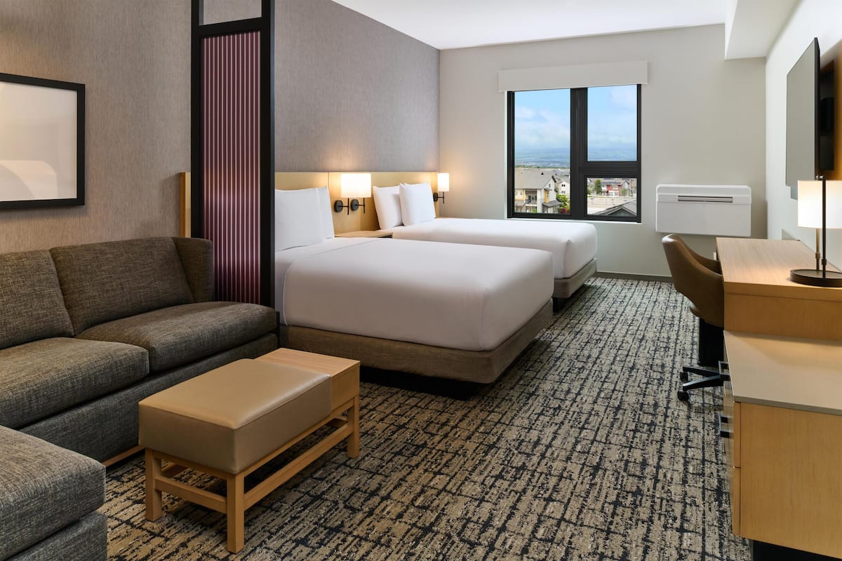 A hotel room with a sofa, two beds, a desk, and a large window with a city view.