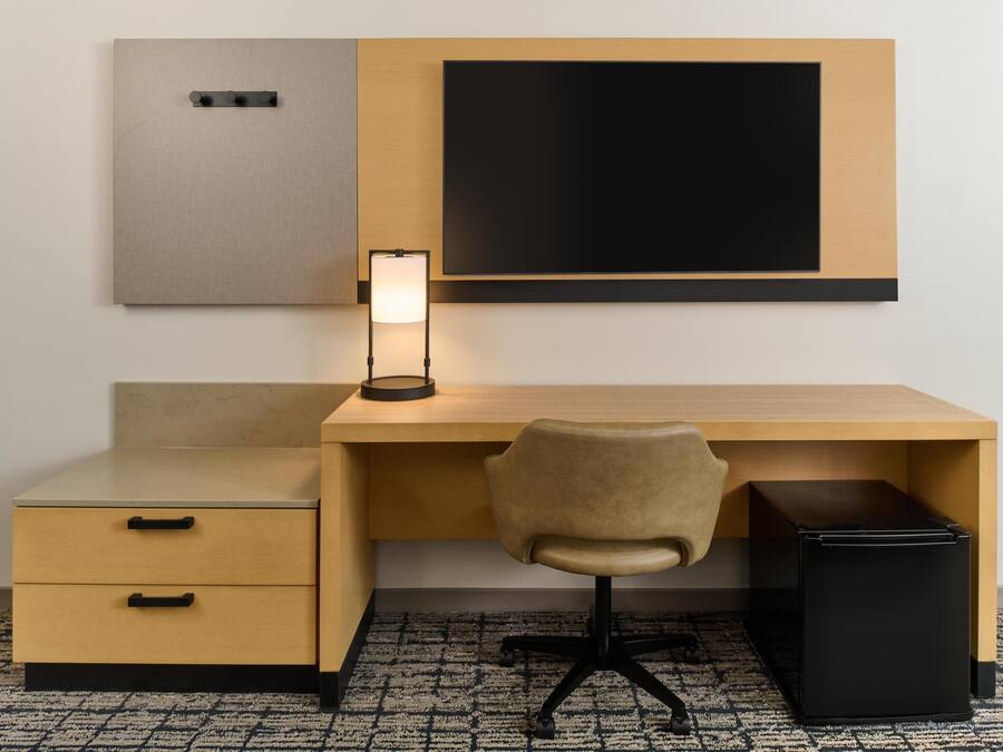 A hotel room with a wooden desk, a chair, a small cabinet, a black trash bin, and a wall-mounted TV.