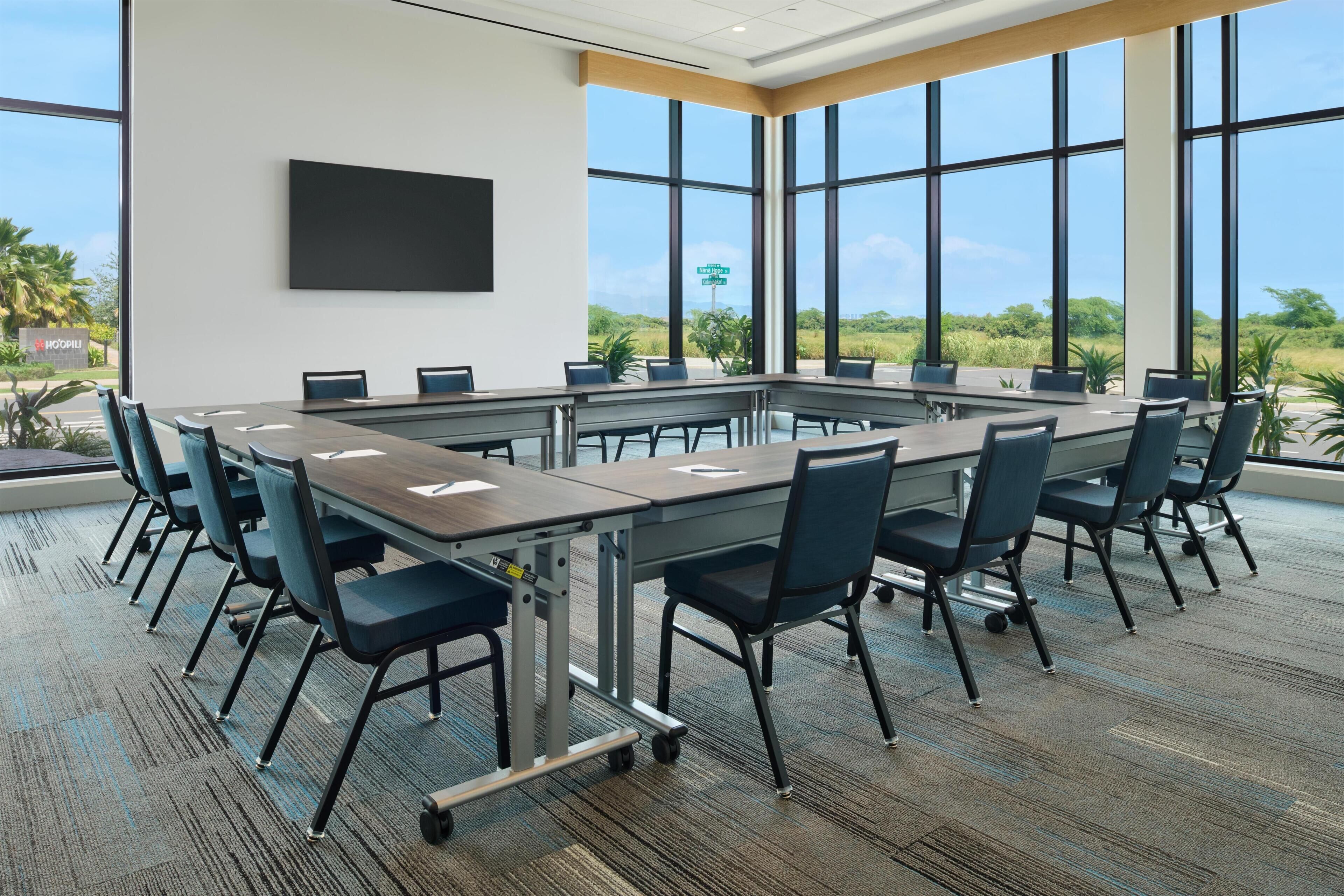 A conference room with a long table, chairs, a flat-screen TV, and large windows with a view of greenery.