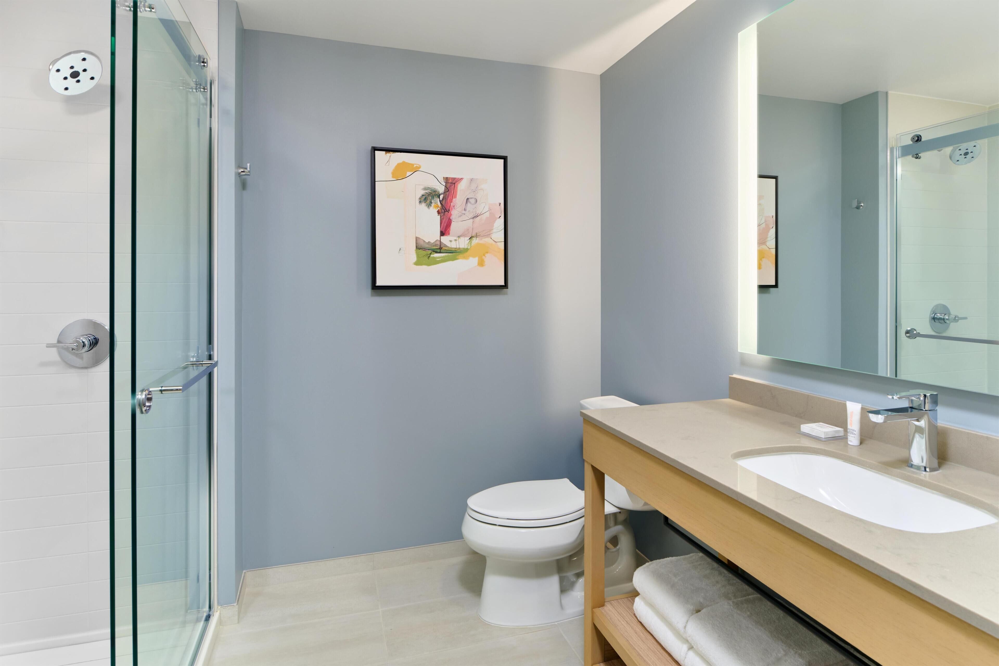 Un baño moderno con cabina de ducha de vidrio, inodoro, lavamanos con espejo y una obra de arte enmarcada en la pared.