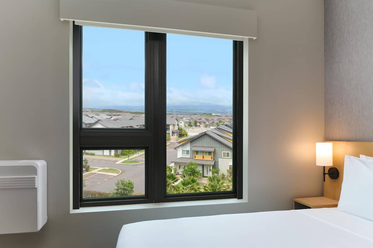 A hotel room with a large window, a bed, a nightstand, and a wall-mounted lamp.