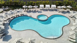 Une grande piscine en forme de rein, entourée de chaises longues et de parasols, avec une terrasse dallée et une clôture délimitant l’espace.