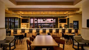 Un bar moderne doté d’un long comptoir, de nombreux espaces pour s’asseoir et d’une vue sur une piscine et des palmiers.