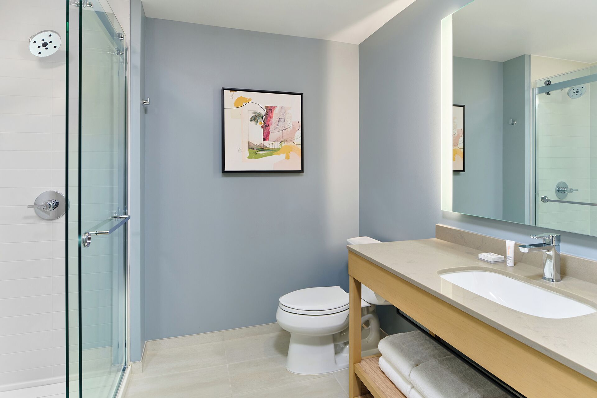 Un baño moderno con cabina de ducha de vidrio, inodoro blanco, tocador beige con espejo y un cuadro abstracto enmarcado en la pared.