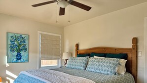 Quarto com estrutura de cama de madeira, ventilador de teto, janela com persianas, luminária de cabeceira e pintura de flores.