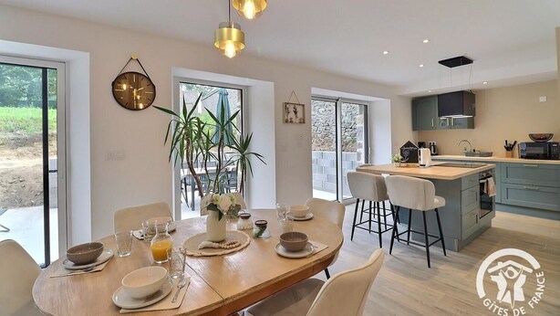 A modern kitchen with a central island, a dining area with a wooden table and chairs, and a large window providing a view of the outdoors. - Le refuge du Mesnil - MESNIL-ROC'H (MESNIL-ROC'H)
