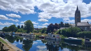 A calm river with boats docked, a church with a tall tower, and a clear blue sky. - Le refuge du Mesnil - MESNIL-ROC'H (MESNIL-ROC'H)