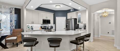 A modern kitchen with a central island, bar stools, and a dining area with a wicker chair.