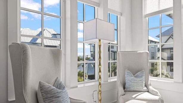 Une pièce dotée de deux fauteuils à haut dossier, d’un lampadaire et de grandes fenêtres donnant sur les maisons voisines.