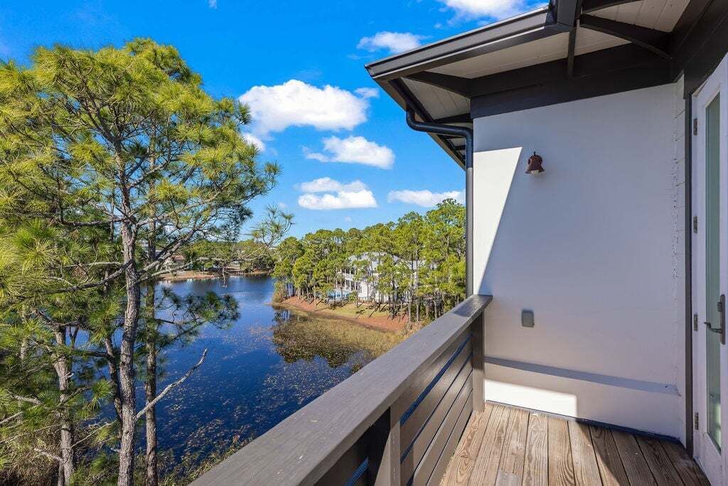 A wooden deck overlooking a calm lake with trees and a white building in the background.
