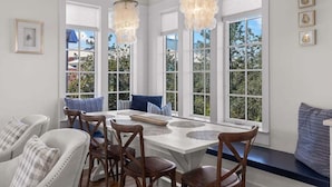 Un espace repas avec une table et des chaises blanches, de grandes fenêtres et des lustres décoratifs.