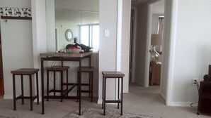 A living room with a white table, a brown bag, and a pair of headphones. There are three wooden stools and a small white table. A kitchen area is visible in the background with a sink and a mirror. - Direct Oceanfront Balcony, 23rd Floor, Peck Plaza Building, Daytona Beach Shores (Daytona Beach Shores)