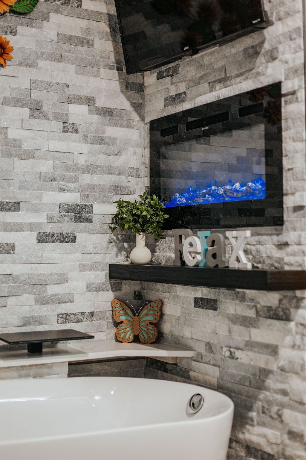 Ein Badezimmer mit Steinwand, einem angebrachten Fernseher, einer weißen Badewanne und Dekoelementen, darunter eine Pflanze und eine Schmetterlingsfigur.