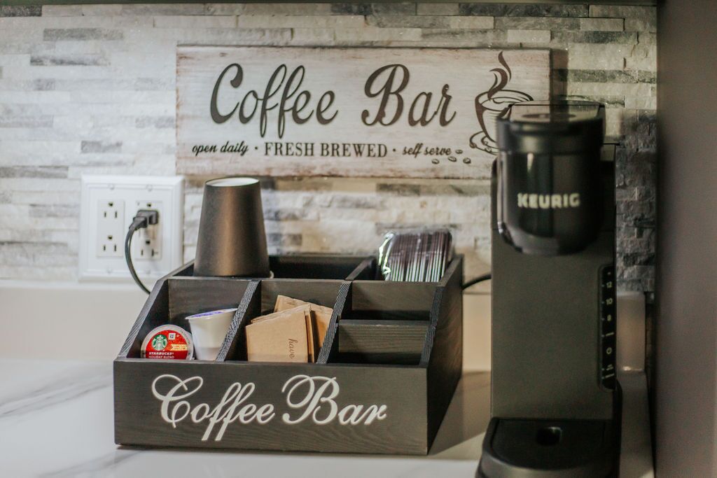 Una estación de café con una máquina Keurig, un molinillo de café y un letrero que dice 'Coffee Bar abierta todos los días, café recién hecho, autoservicio'.