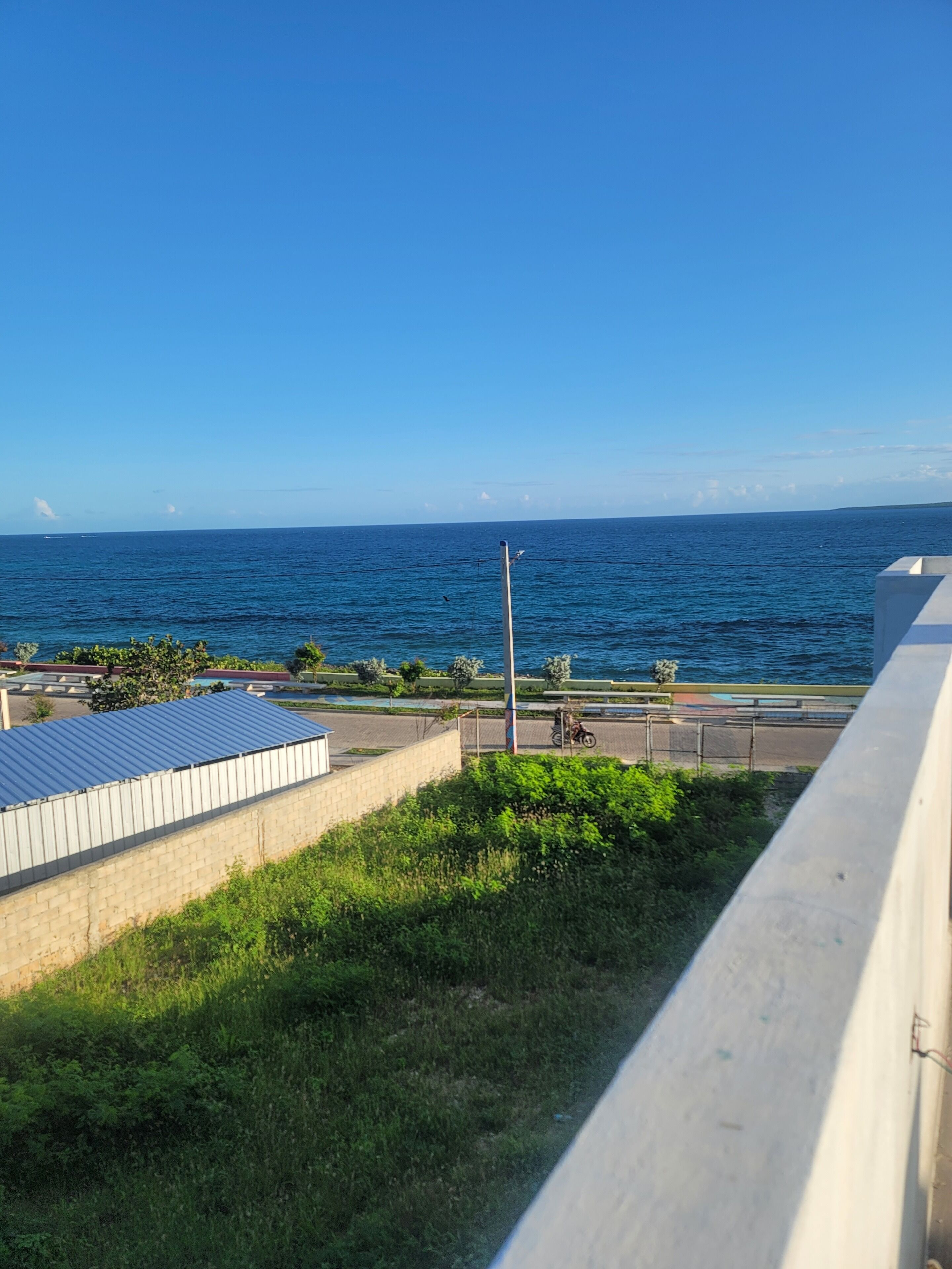 A coastal view with a clear sky, a road with a parked motorcycle, and a building with a blue roof.