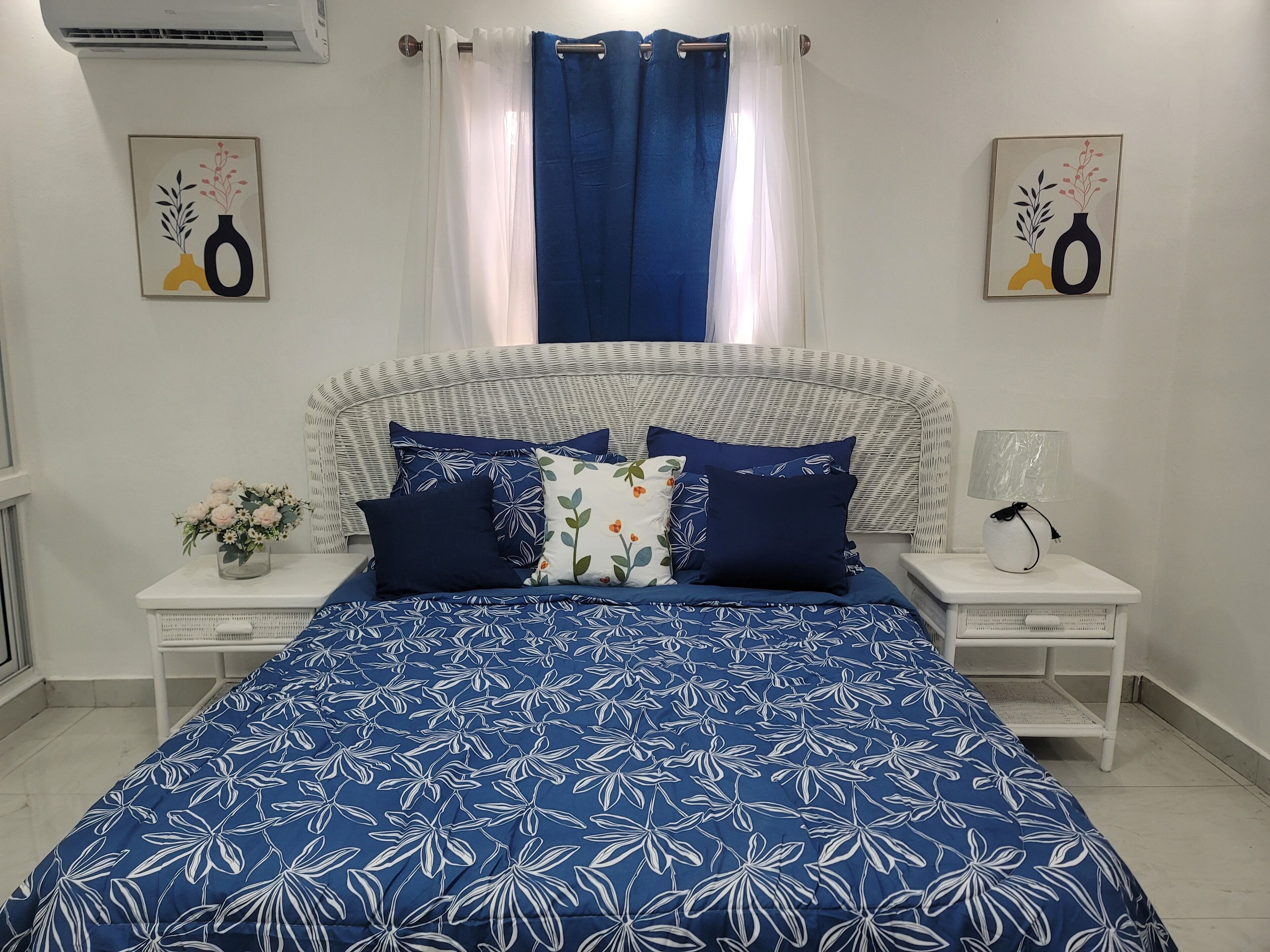Een slaapkamer met een groot bed, witte nachtkastjes, een bedlampje en een raam met gordijnen.