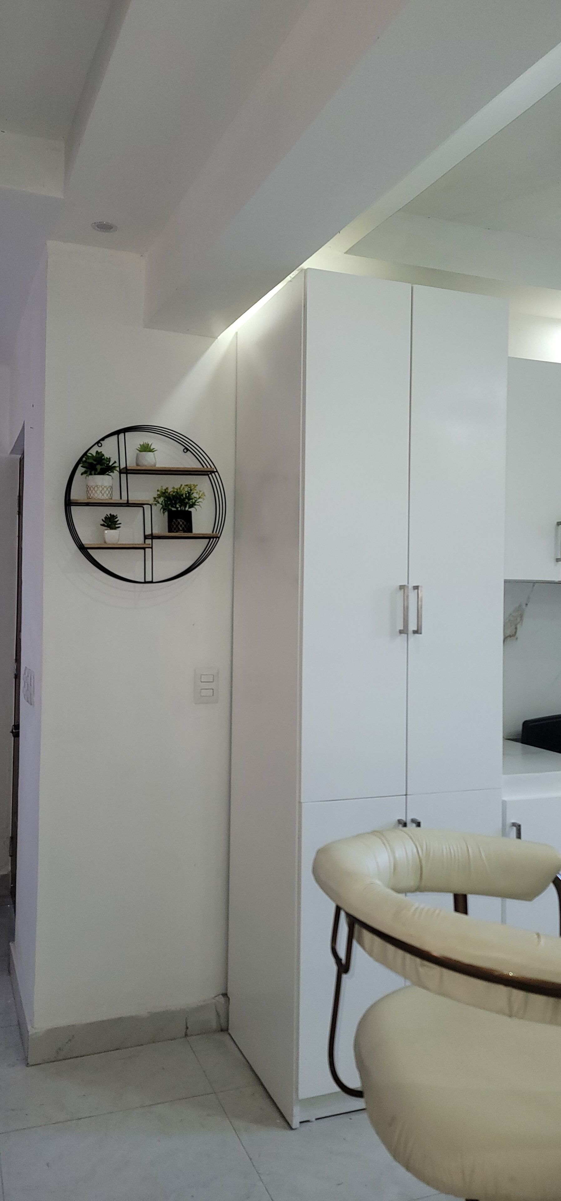 Una habitación moderna con un armario blanco, un estante circular con plantas y una silla color beige.