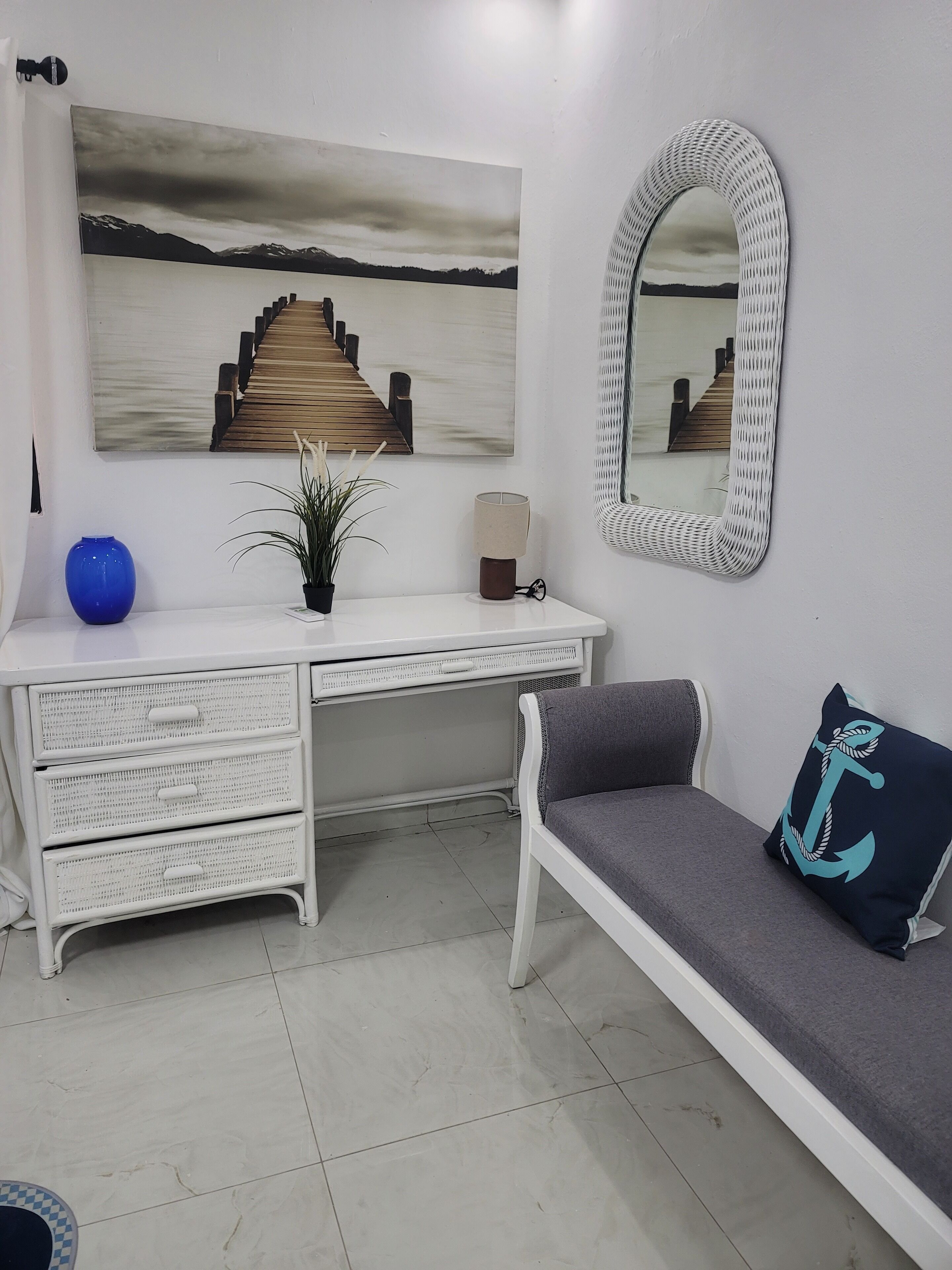 Quarto com escrivaninha branca, vaso azul, planta, luminária, espelho e sofá cinza com almofada azul. Há também um quadro na parede retratando uma escada de madeira.