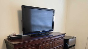 A wooden dresser with a flat-screen TV on top, a small trash bin, and a power outlet on the wall.