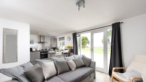 A modern living room with a grey sectional sofa, a wooden chair, and a dining area with a table and chairs. - Wheal Prosper, Wheal Dream - Hot Tub - Free Parking & WiFi - Pet Friendly (Helston)