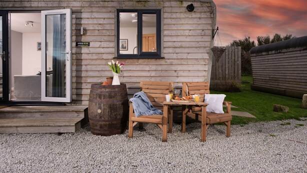 A rustic wooden cabin with a glass door entrance, a small table with breakfast items, and a bench. - Wheal Prosper, Wheal Dream - Hot Tub - Free Parking & WiFi - Pet Friendly (Helston)