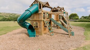 A wooden play structure with slides and climbing elements, set on a gravel surface with a grassy field in the background. - Wheal Prosper, Wheal Dream - Hot Tub - Free Parking & WiFi - Pet Friendly (Helston)